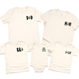 Puede incluir: Se muestran cinco camisetas de color crema y un body. Cada camisa tiene un gr&aacute;fico en negro y verde. Las camisas dicen "DAD", "MOM", "BR" y "SASSY". El body dice "ONE HAPPY CAMPER".