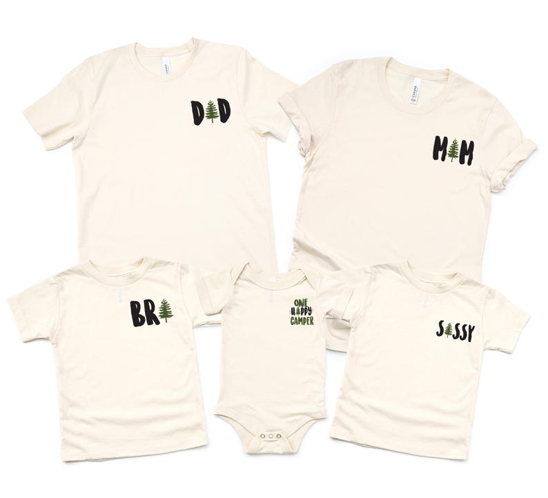 Puede incluir: Se muestran cinco camisetas de color crema y un body. Cada camisa tiene un gr&aacute;fico en negro y verde. Las camisas dicen "DAD", "MOM", "BR" y "SASSY". El body dice "ONE HAPPY CAMPER".