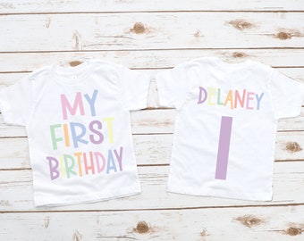 May include: Two white t-shirts with pastel text. One shirt says "MY FIRST BIRTHDAY" in colorful letters. The other shirt has the name "DELANEY" and the number "1" in pastel colors. The shirts are on a light wood background.