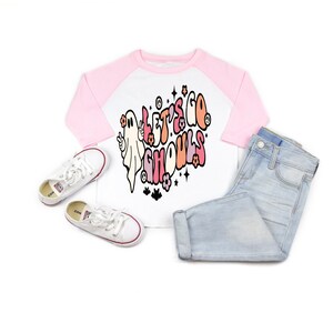 May include: A white and pink raglan-style shirt with the text "LET'S GO GHOULS" and a ghost graphic. The shirt is paired with light blue jeans and white sneakers with red trim. The outfit is displayed on a white background.