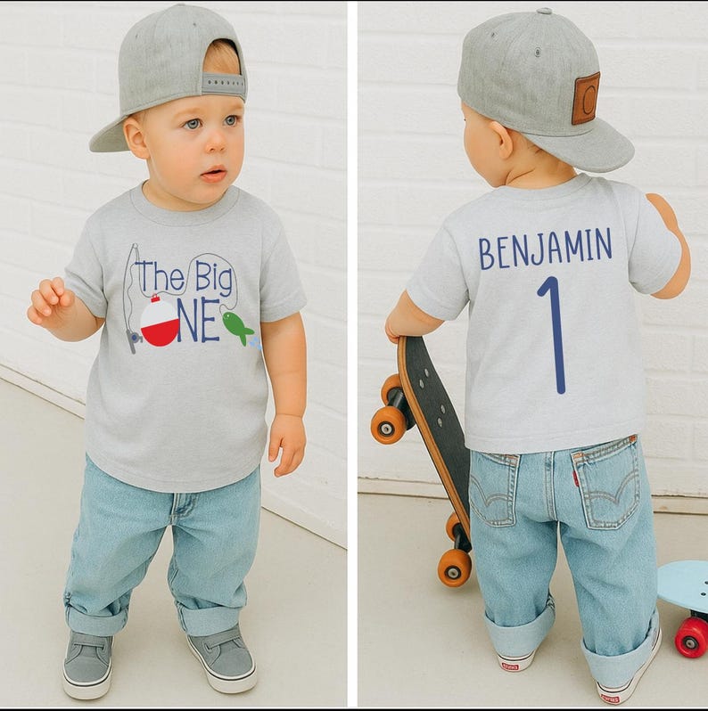 Op de afbeelding: Lichtgrijs T-shirt met de tekst "The Big One" en een vissen-thema afbeelding. De achterkant van het shirt heeft de naam "BENJAMIN" en het nummer "1". Het kind draagt een lichtblauwe jeans en een grijze baseball cap.