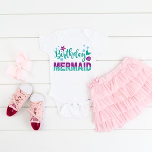 May include: White bodysuit with the text "Birthday Mermaid" in purple and green with a mermaid tail, starfish, and bubbles. The bodysuit is laid out on a white wooden surface with a pink tutu, a pink bow, and pink sneakers.