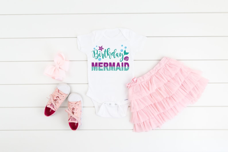 May include: White bodysuit with the text "Birthday Mermaid" in purple and green with a mermaid tail, starfish, and bubbles. The bodysuit is laid out on a white wooden surface with a pink tutu, a pink bow, and pink sneakers.