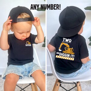 May include: A black t-shirt featuring a yellow excavator graphic and the text "HAYDEN" on the front, and a larger yellow excavator graphic with the text "TWO AND DIGGIN IT!" on the back, paired with a plain black baseball cap. The text "ANY NUMBER!" appears.