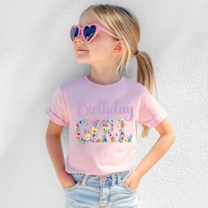 May include: A light pink short-sleeve t-shirt featuring the text Birthday Girl in a purple script font above the word GIRL in a large, bold, floral-patterned print with various colorful flowers, paired with light blue denim jeans and heart-shaped sunglasses.