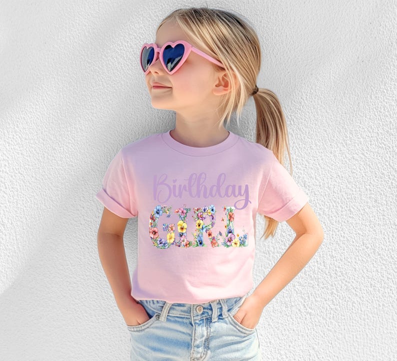 May include: A light pink short-sleeve t-shirt featuring the text Birthday Girl in a purple script font above the word GIRL in a large, bold, floral-patterned print with various colorful flowers, paired with light blue denim jeans and heart-shaped sunglasses.