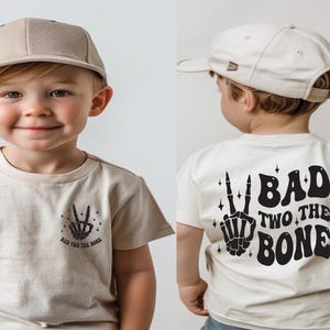 Puede incluir: Un ni&ntilde;o peque&ntilde;o con una camiseta beige con un gr&aacute;fico de mano de esqueleto negro y el texto "Bad Two The Bone". El ni&ntilde;o tambi&eacute;n lleva una gorra de b&eacute;isbol beige.
