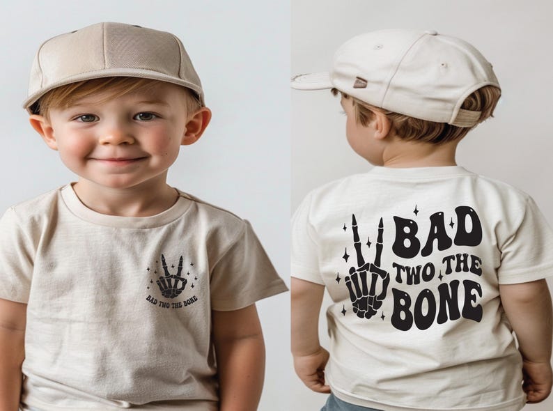 Puede incluir: Un ni&ntilde;o peque&ntilde;o con una camiseta beige con un gr&aacute;fico de mano de esqueleto negro y el texto "Bad Two The Bone". El ni&ntilde;o tambi&eacute;n lleva una gorra de b&eacute;isbol beige.