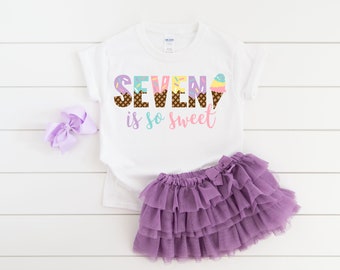 May include: White t-shirt with the text "SEVEN is so sweet" in colorful letters decorated with ice cream cones. The shirt is paired with a purple tulle skirt.