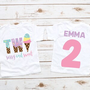 Pu&ograve; includere: Due magliette bianche con un design a forma di cono gelato. La maglietta anteriore dice "TWO sassy and sweet" con un cono gelato. La maglietta posteriore dice "EMMA 2" in rosa.