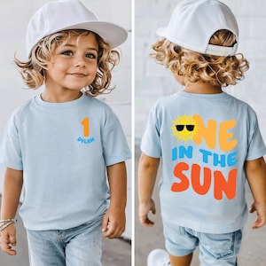 May include: A light blue t-shirt featuring a small orange number one and the name Dylan on the front. The back displays a yellow sun graphic with black sunglasses and the text ONE IN THE SUN in large orange and blue lettering.