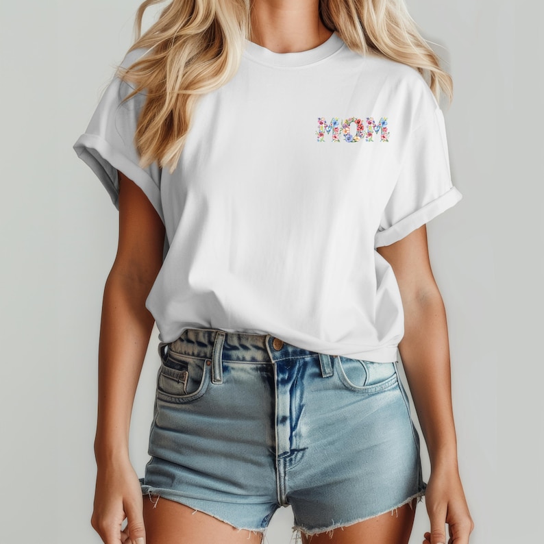 May include: A white short-sleeved t-shirt with a crew neckline and rolled sleeves. The chest features the word MOM printed in a colorful floral pattern consisting of small blue, pink, and red flowers with green leaves and stems.
