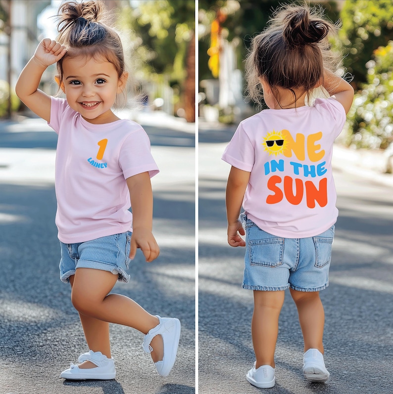 May include: A light pink short-sleeved t-shirt featuring the number 1 and the name Lainey on the front chest. The back of the shirt displays a yellow sun graphic with sunglasses and the text ONE IN THE SUN in blue and orange lettering.