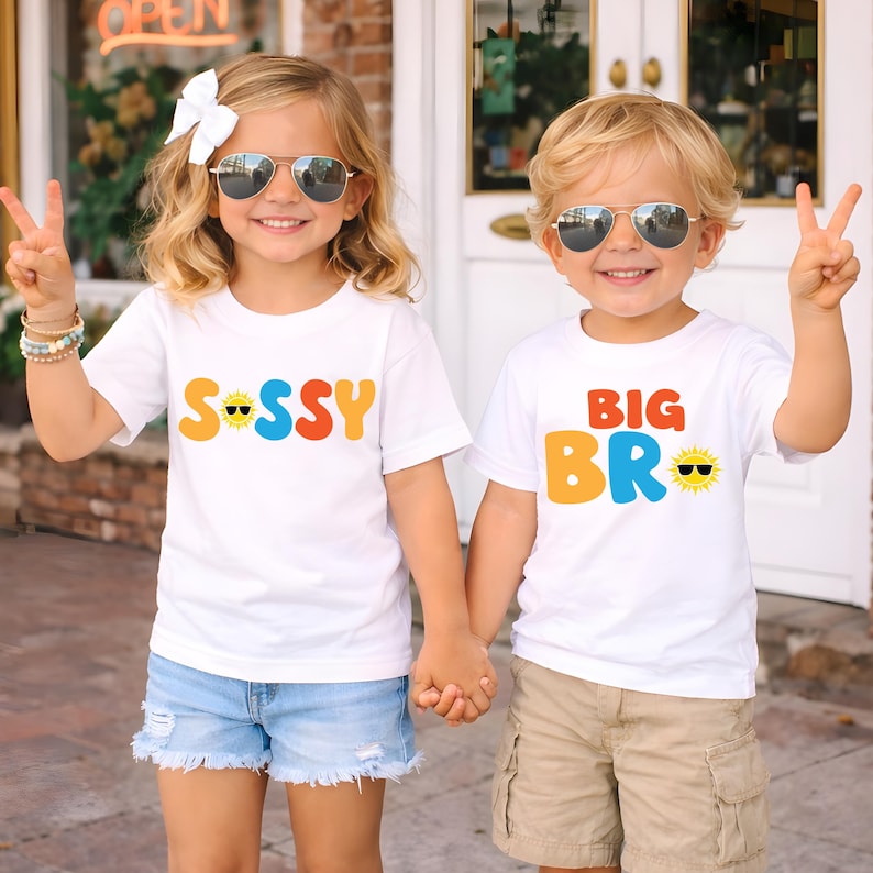 May include: Two white short-sleeved t-shirts featuring colourful graphic text. One shirt displays the word SISSY with a sun icon wearing sunglasses, and the other displays BIG BRO with a sun icon wearing sunglasses. Both shirts have crew necks.