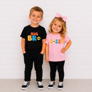 May include: A black t-shirt with the text BIG BRO in orange and blue with a yellow sun icon wearing sunglasses, and a light pink t-shirt with the text S-SSY in yellow, pink, and orange with a yellow smiley face icon wearing sunglasses.