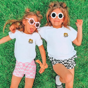 May include: Two white short-sleeve t-shirts featuring a small graphic on the left chest of a yellow construction dump truck above the text SISSY. The shirts are paired with checkered shorts in pink and white or black and white patterns.