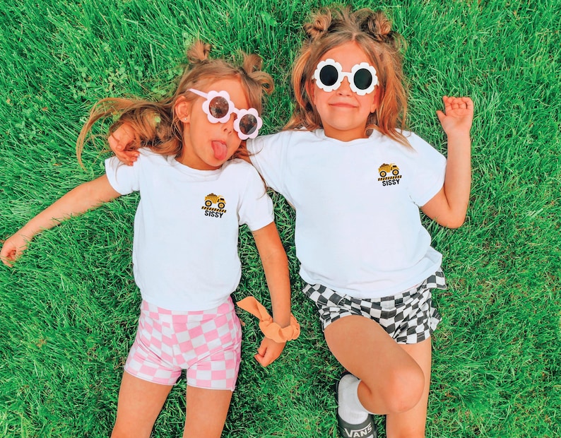 May include: Two white short-sleeve t-shirts featuring a small graphic on the left chest of a yellow construction dump truck above the text SISSY. The shirts are paired with checkered shorts in pink and white or black and white patterns.