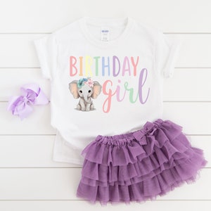 May include: White t-shirt with a rainbow colored "Birthday girl" text and a cartoon elephant wearing a flower crown. The shirt is paired with a purple tulle skirt.