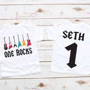 May include: Two white t-shirts with a rock and roll theme. The front of the shirt has a row of colorful guitars with the text "One Rocks". The back of the shirt has the name "Seth" and the number "1" in a bold black font.