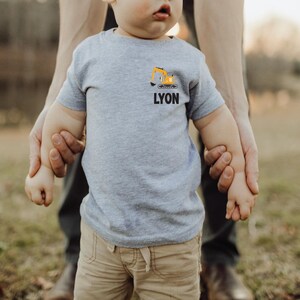 May include: A light gray short-sleeved t-shirt featuring a small graphic of a yellow excavator above the name LYON printed in bold black capital letters on the left chest area of the garment.