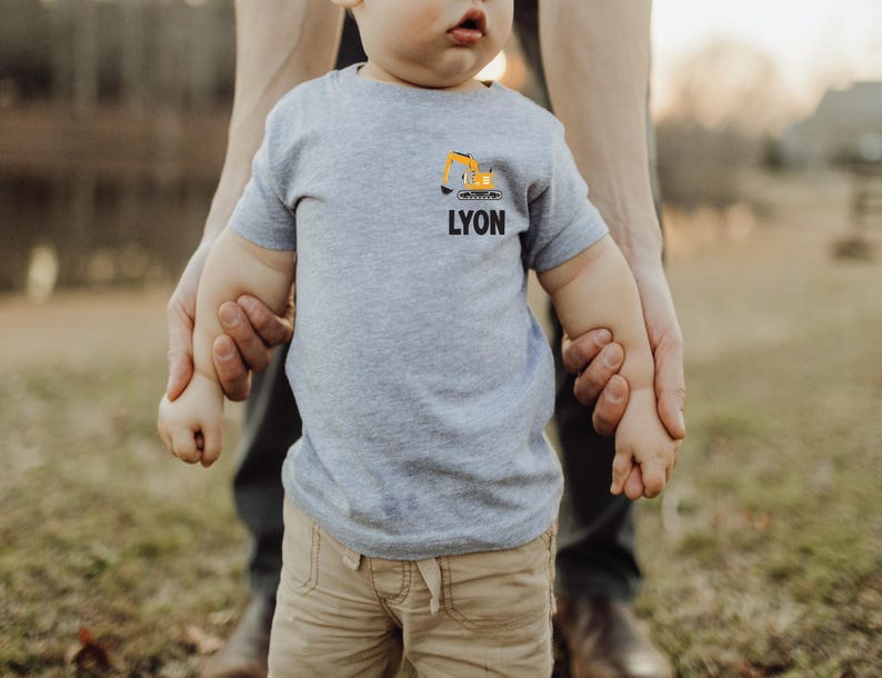 May include: A light gray short-sleeved t-shirt featuring a small graphic of a yellow excavator above the name LYON printed in bold black capital letters on the left chest area of the garment.