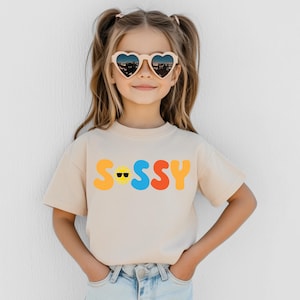 May include: A beige short-sleeve t-shirt featuring the word SASSY printed in large, colourful, rounded letters across the chest. The letter A is replaced by a yellow sun icon wearing black sunglasses. The shirt has a crew neckline.