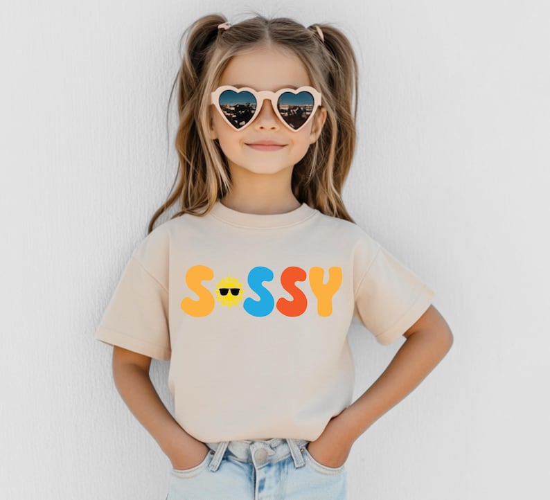 May include: A beige short-sleeve t-shirt featuring the word SASSY printed in large, colourful, rounded letters across the chest. The letter A is replaced by a yellow sun icon wearing black sunglasses. The shirt has a crew neckline.