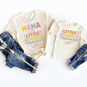 May include: Two cream-colored t-shirts and two pairs of blue denim jeans. The larger shirt features the text MAMA of the LITTLE MISS THREENAGER, and the smaller shirt features LITTLE MISS THREENAGER, both decorated with colorful floral graphics.