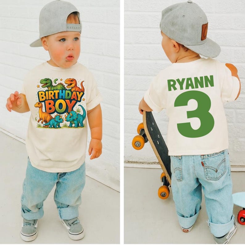 May include: A cream-colored t-shirt with a colorful dinosaur graphic and the words "BIRTHDAY BOY." The back of the shirt has the name "RYANN" and the number "3" in green. The child is wearing light blue jeans and a gray baseball cap.