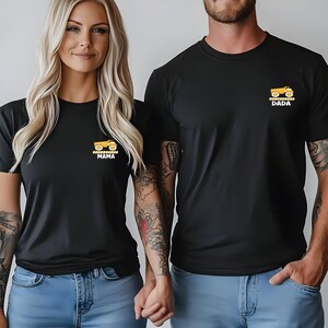 May include: Two black crew neck t-shirts featuring a small graphic on the left chest. Each shirt displays a yellow dump truck icon above a yellow and black striped caution bar. One shirt has the word MAMA printed below, and the other has DADA.