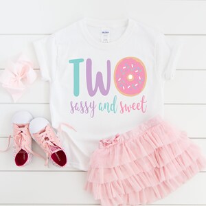 May include: White t-shirt with the text "TWO sassy and sweet" and a pink donut with sprinkles. The shirt is paired with a pink tulle skirt and pink sneakers.