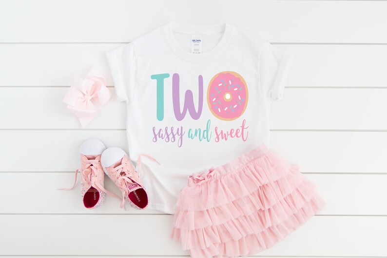 May include: White t-shirt with the text "TWO sassy and sweet" and a pink donut with sprinkles. The shirt is paired with a pink tulle skirt and pink sneakers.