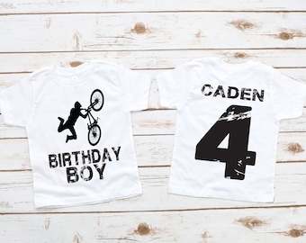 May include: Two white t-shirts with a black graphic design. The front of one shirt features a silhouette of a person doing a bike trick with the text "Birthday Boy". The back of the other shirt has the name "CADEN" and the number "4" in black.