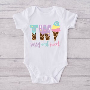 Pu&ograve; includere: Body bianco per neonato con il numero due scritto usando coni gelato. I coni gelato sono decorati con codette e il testo dice "sassy and sweet".
