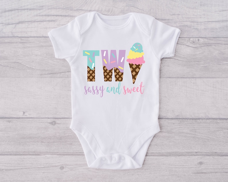 Pu&ograve; includere: Body bianco per neonato con il numero due scritto usando coni gelato. I coni gelato sono decorati con codette e il testo dice "sassy and sweet".