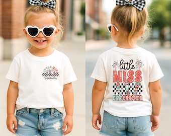 Puede incluir: Una camiseta blanca con un gráfico rojo y azul de fuegos artificiales y el texto "little miss firecracker". La camiseta la lleva puesta un niño pequeño con el pelo rubio y un lazo blanco y negro.