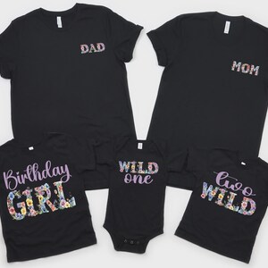 May include: A set of five black garments including two adult t-shirts with floral DAD and MOM text, a child t-shirt with Birthday GIRL text, a baby bodysuit with WILD one text, and a child t-shirt with two WILD text in floral patterns.