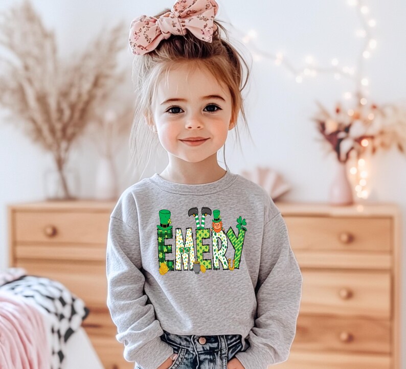 May include: A gray sweatshirt with a green, yellow, and orange design of leprechauns spelling out the name "EMERY".