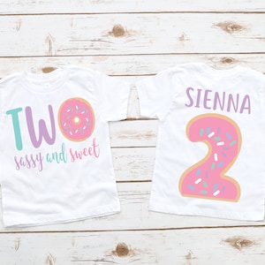 May include: Two white t-shirts with a donut design. One shirt says "TWO sassy and sweet" with a pink donut with sprinkles. The other shirt has a pink donut shaped number "2" with sprinkles and the name "SIENNA" above it.