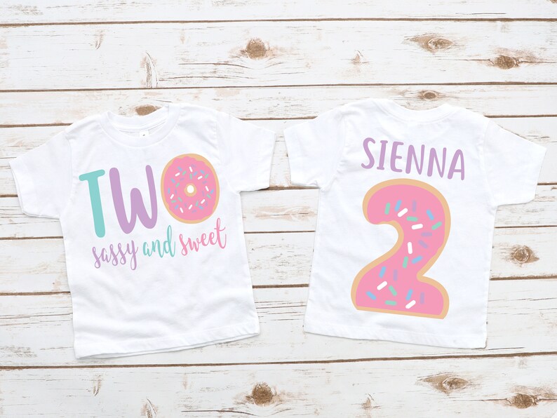 May include: Two white t-shirts with a donut design. One shirt says "TWO sassy and sweet" with a pink donut with sprinkles. The other shirt has a pink donut shaped number "2" with sprinkles and the name "SIENNA" above it.