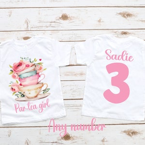 May include: White t-shirt with a stack of teacups and flowers printed on the front. The back of the shirt has the name "Sadie" and the number "3" printed in pink.