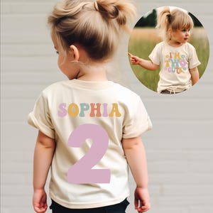 May include: A cream colored t-shirt with the name "Sophia" in colorful letters and the number "2" in pink. The shirt is being worn by a child.