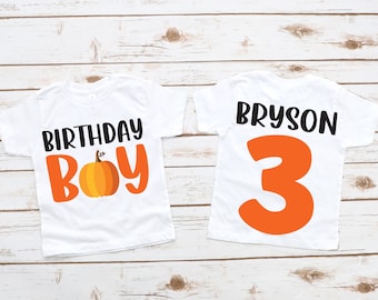 Può includere: Due magliette bianche con testo arancione e nero. Una maglietta dice "BIRTHDAY BOY" con una grafica di zucca. L'altra maglietta dice "BRYSON" e il numero "3".