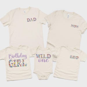 以下が含まれることがあります： クリーム色の衣類5点セット。大人用Tシャツ2枚（花柄のDADとMOMの文字入り）、子供用Tシャツ1枚（紫と花柄のBirthday GIRLの文字入り）、ベビー用ボディスーツ1枚（紫と花柄のWILD oneの文字入り）、子供用Tシャツ1枚（花柄のBROの文字入り）が含まれています。