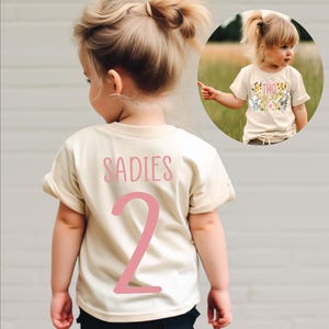 May include: A light beige t-shirt with pink text that says "SADIES 2" on the back. The shirt is being worn by a child.