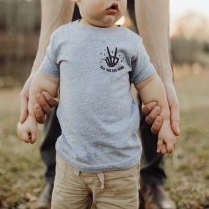 Puede incluir: Un ni&ntilde;o peque&ntilde;o con una camiseta gris con un gr&aacute;fico de signo de paz negro y el texto "Rad This Good" est&aacute; siendo sostenido por un adulto. El ni&ntilde;o lleva pantalones caqui.