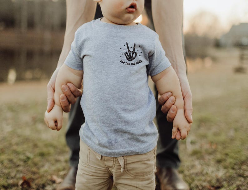Puede incluir: Un ni&ntilde;o peque&ntilde;o con una camiseta gris con un gr&aacute;fico de signo de paz negro y el texto "Rad This Good" est&aacute; siendo sostenido por un adulto. El ni&ntilde;o lleva pantalones caqui.