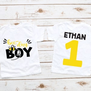 May include: Two white t-shirts with a bee-themed design. The front of one shirt says "bee-day boy" with a cartoon bee and the back of the other shirt says "ETHAN" with the number "1" in yellow.