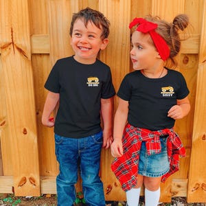 May include: Two black short-sleeve t-shirts featuring a small yellow dump truck graphic on the left chest. The left shirt has the text BIG BRO below the truck, and the right shirt has the text SISSY below the truck, both in white lettering.