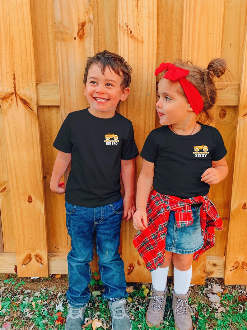 May include: Two black short-sleeve t-shirts featuring a small yellow dump truck graphic on the left chest. The left shirt has the text BIG BRO below the truck, and the right shirt has the text SISSY below the truck, both in white lettering.